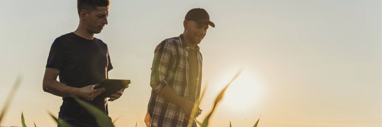 Due persone camminano in un campo coltivato al tramonto: una consulta un tablet e l’altra indossa una camicia a quadri e cappellino