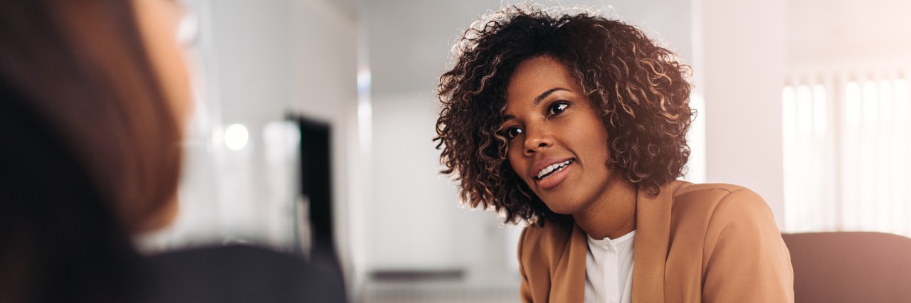 a woman in a brown blazer listens intently to something another woman is saying.