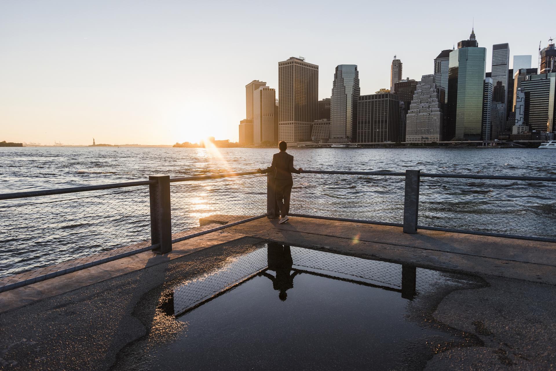 man in suit standing at the waters edge looking at the city in the distance while the sun is rising