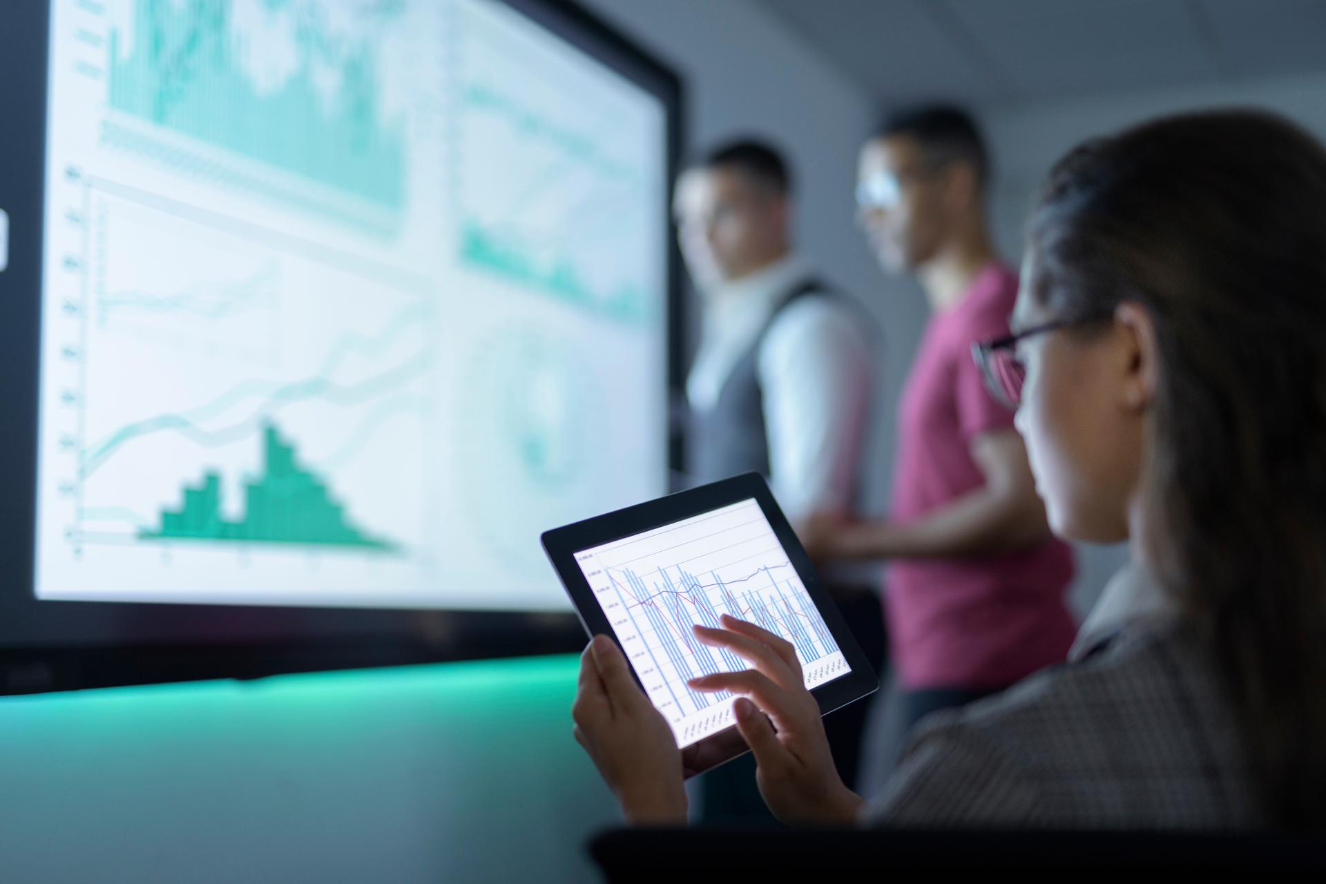 woman on tablet controling smartboard in background with two males looking at board with metrics on it