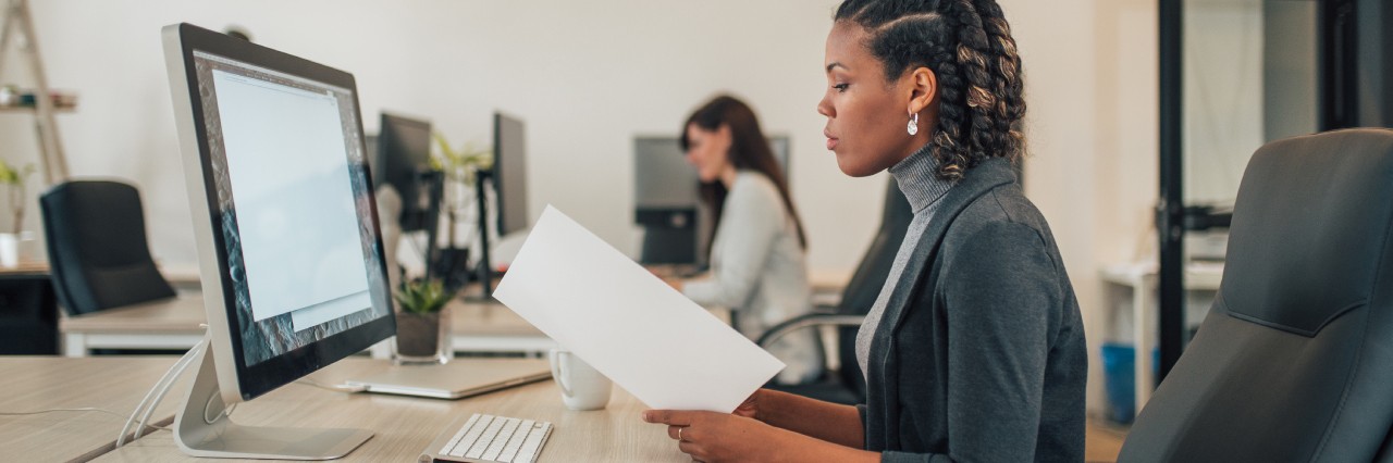 Lady holding a white brochure seemingly double checking with the information on her computer screen sitting at a desk in a office.