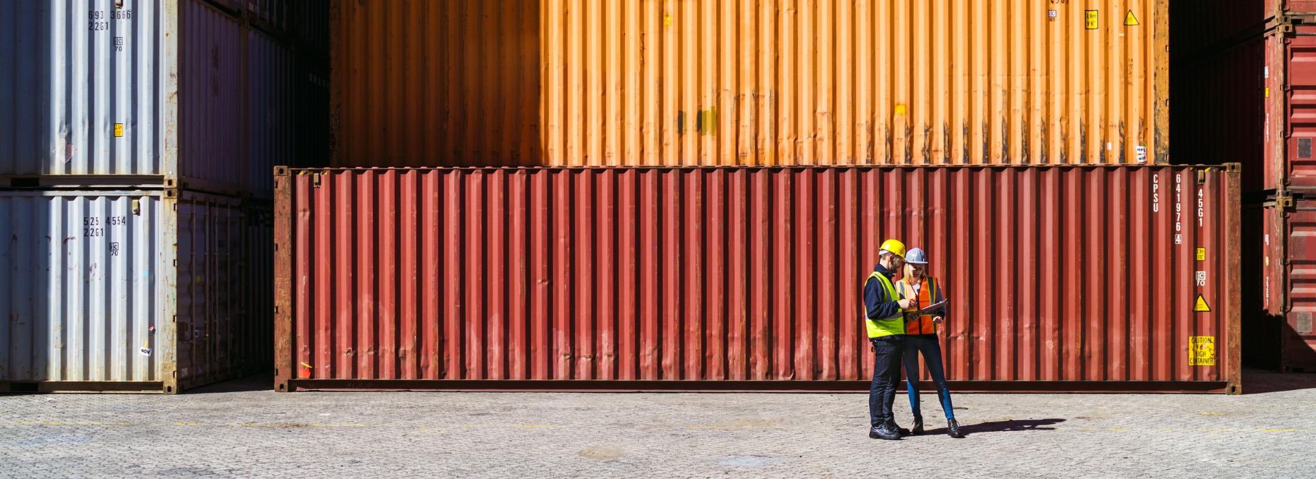 Due operai con caschi protettivi e gilet ad alta visibilità controllano dei dati davanti a dei container