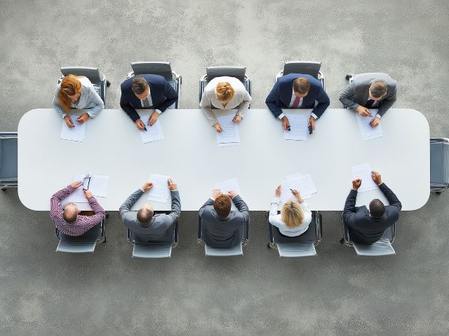 Ripresa dall'alto di un business panel collaborativo durante un meeting raccolto intorno a un grande tavolo per discutere strategie e idee