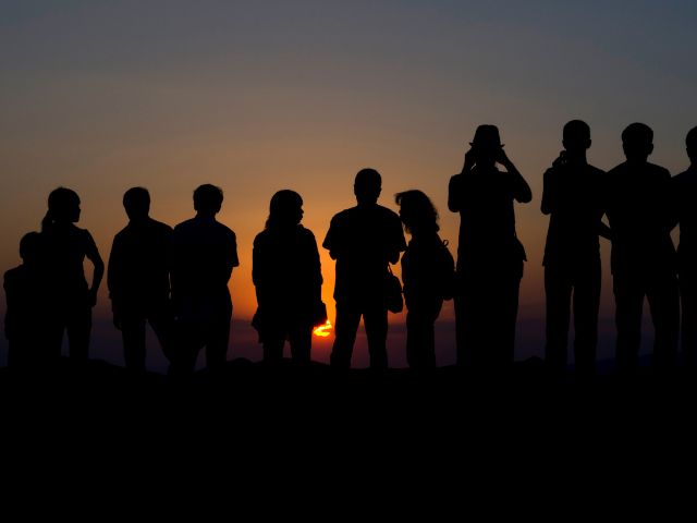 Undici persone di spalle e in penombra, ammirano e scattano foto al tramonto