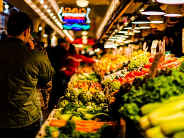 Mercato cittadino con banchi di ortofrutta e persone che fanno la spesa