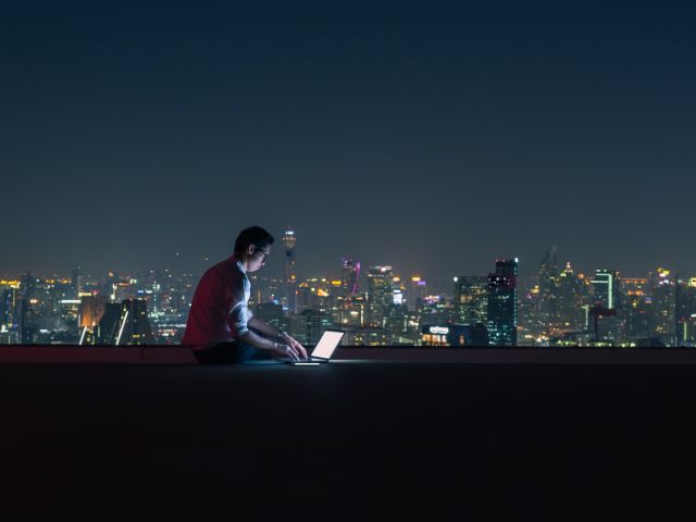 Un imprenditore lavora sul terrazzo di un palazzo, di sera, con vista sulla città illuminata