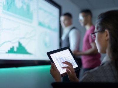 Tre colleghi esaminano dei dati proiettati su un grande schermo e su un tablet